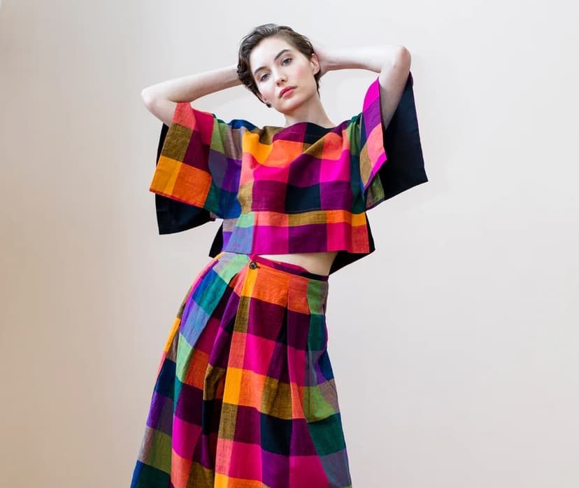 Woman wearing a vibrant, multicolored checkered two-piece set with wide-leg pants and a boxy top, posing confidently with hands behind her head against a neutral background.