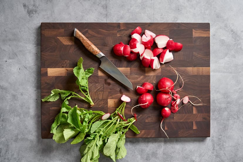 Fresh radishes, some whole and some chopped, with their leafy greens on a dark wood cutting board beside a chef’s knife with a burl wood handle and the brand name Steelport engraved on the blade.