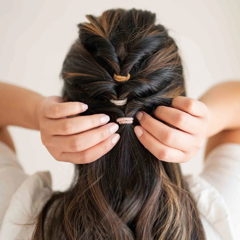 A person styles their dark, wavy hair using three KOOSHOO biodegradable hair ties in earth-toned colors, securing a twisted half-up hairstyle with both hands.