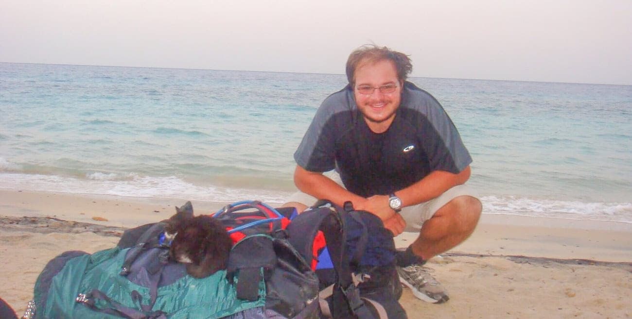 a man on a beach crouched behind two backpacks with a black kitten curled up on one of them