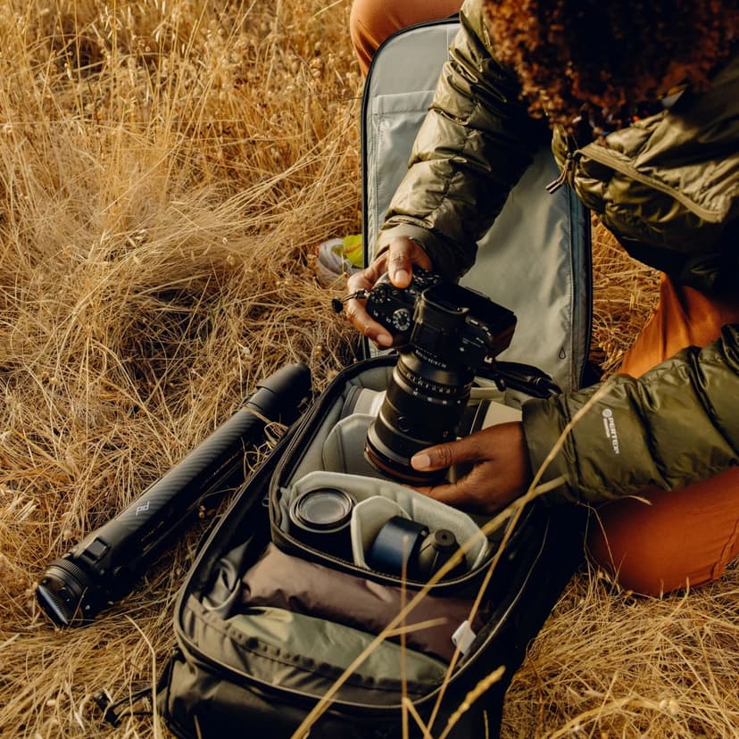 A person in a field crouches down and looks down at the screen of a large digital camera, which is over a Peak Design backpack that is specially packed to accommodate the camera, its lenses, and more.