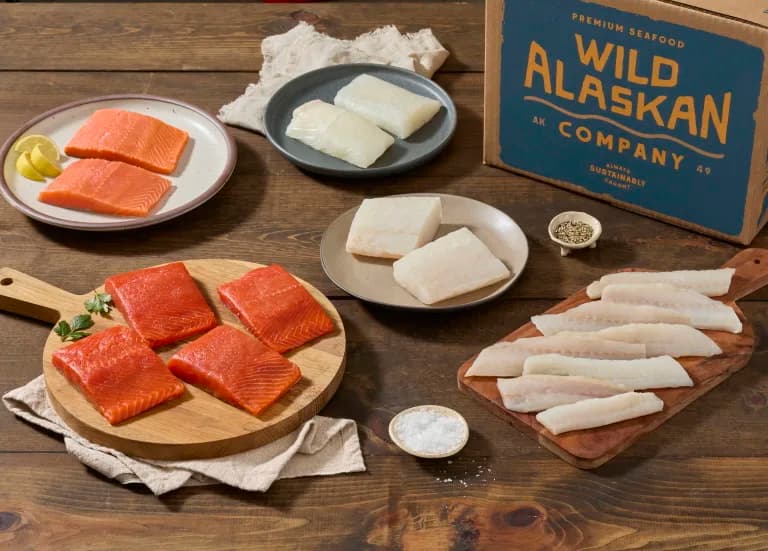 A table with uncooked filets of salmon and white fish on plates next to a carboard box with the words Wild Alaskan Company printed on it.